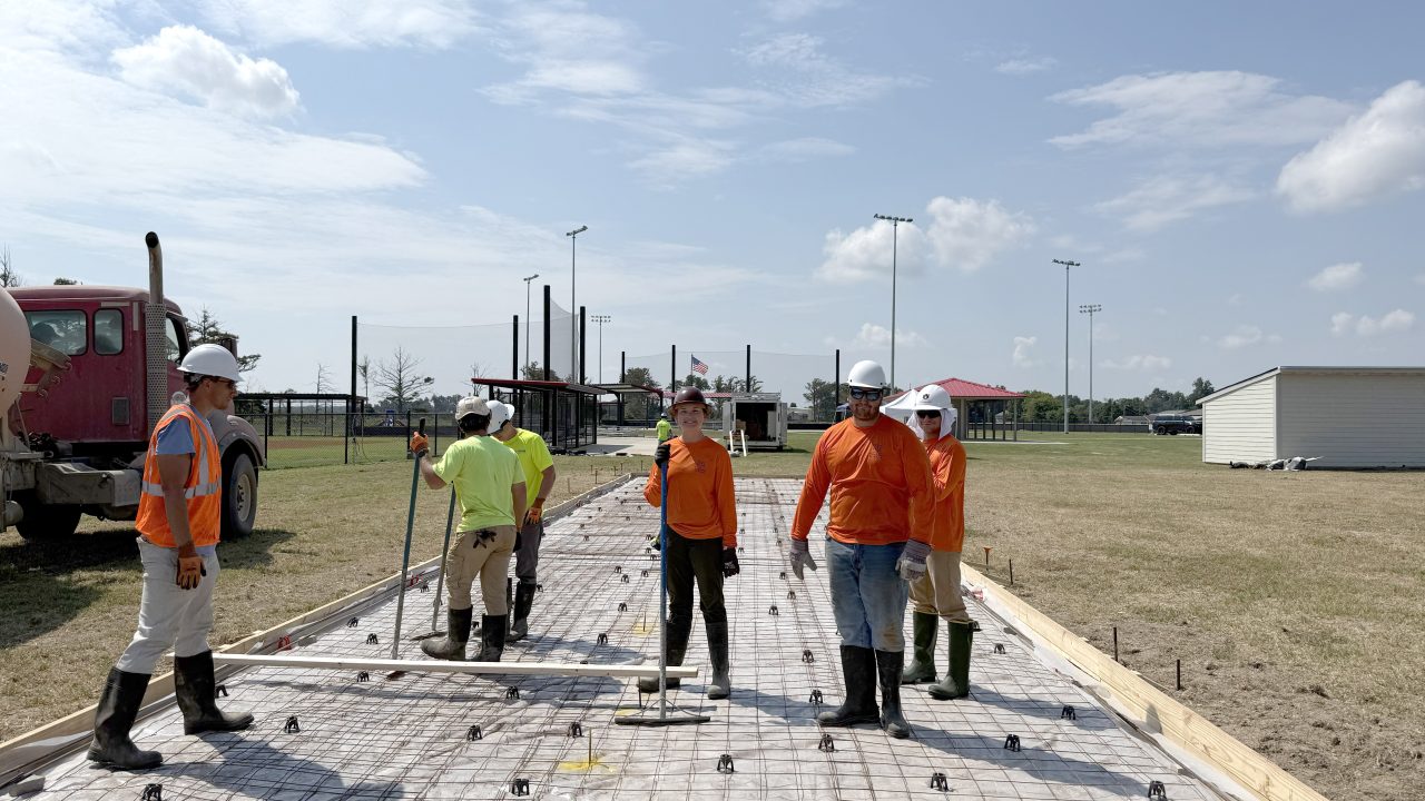 Six construction workers in safety gear stand on a rebar grid, preparing to pour concrete at an outdoor site with sports field lighting in the background.