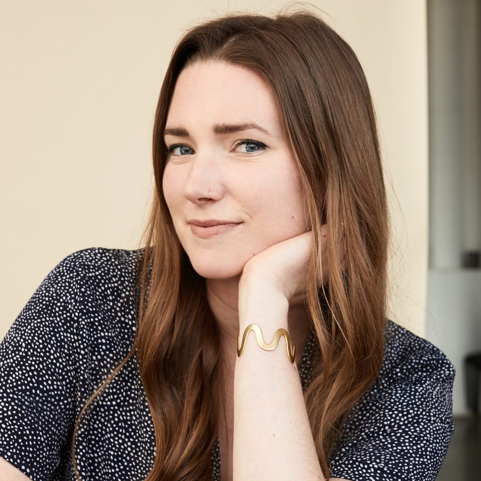 A woman with long brown hair sits indoors, resting her chin on her hand, wearing a patterned short-sleeve top and a gold bracelet.