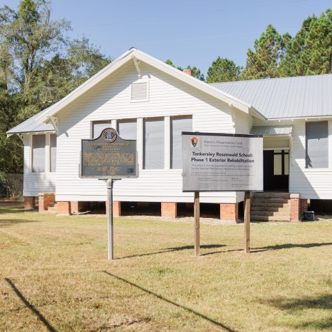 A white, single-story building with a metal roof stands behind two informational signs on a grassy lot, surrounded by trees.