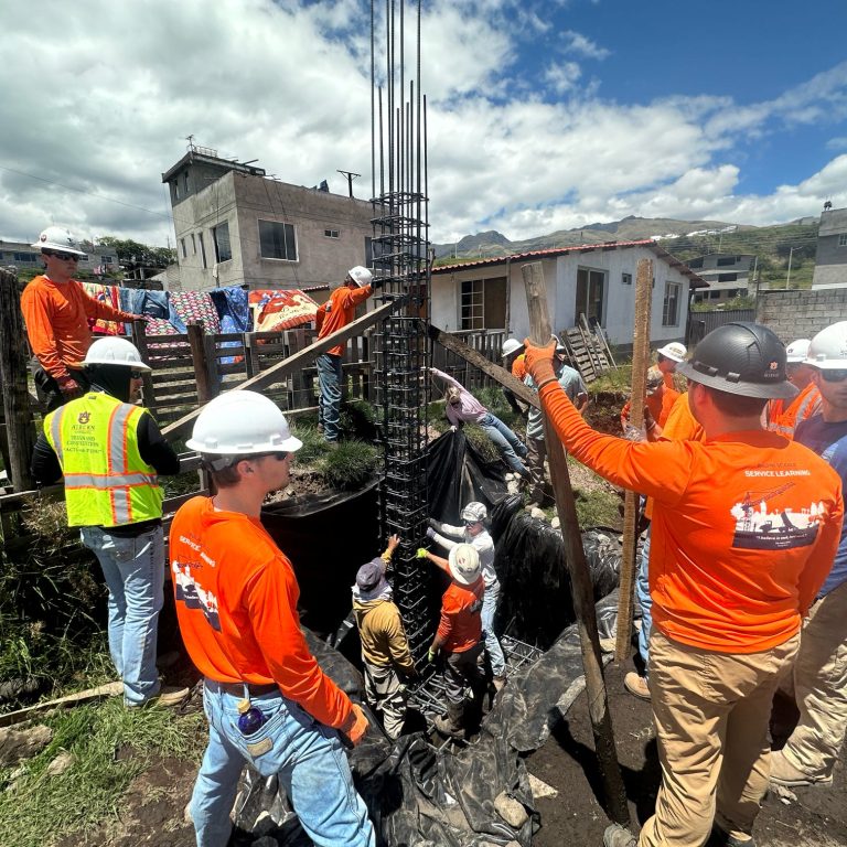 A group of construction workers in orange shirts and white helmets assemble rebar for a building foundation at an outdoor construction site under a partly cloudy sky.