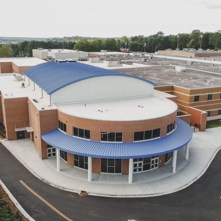 Aerial view of a large, modern school building with a curved blue roof, brick exterior, and adjacent parking lot surrounded by trees.