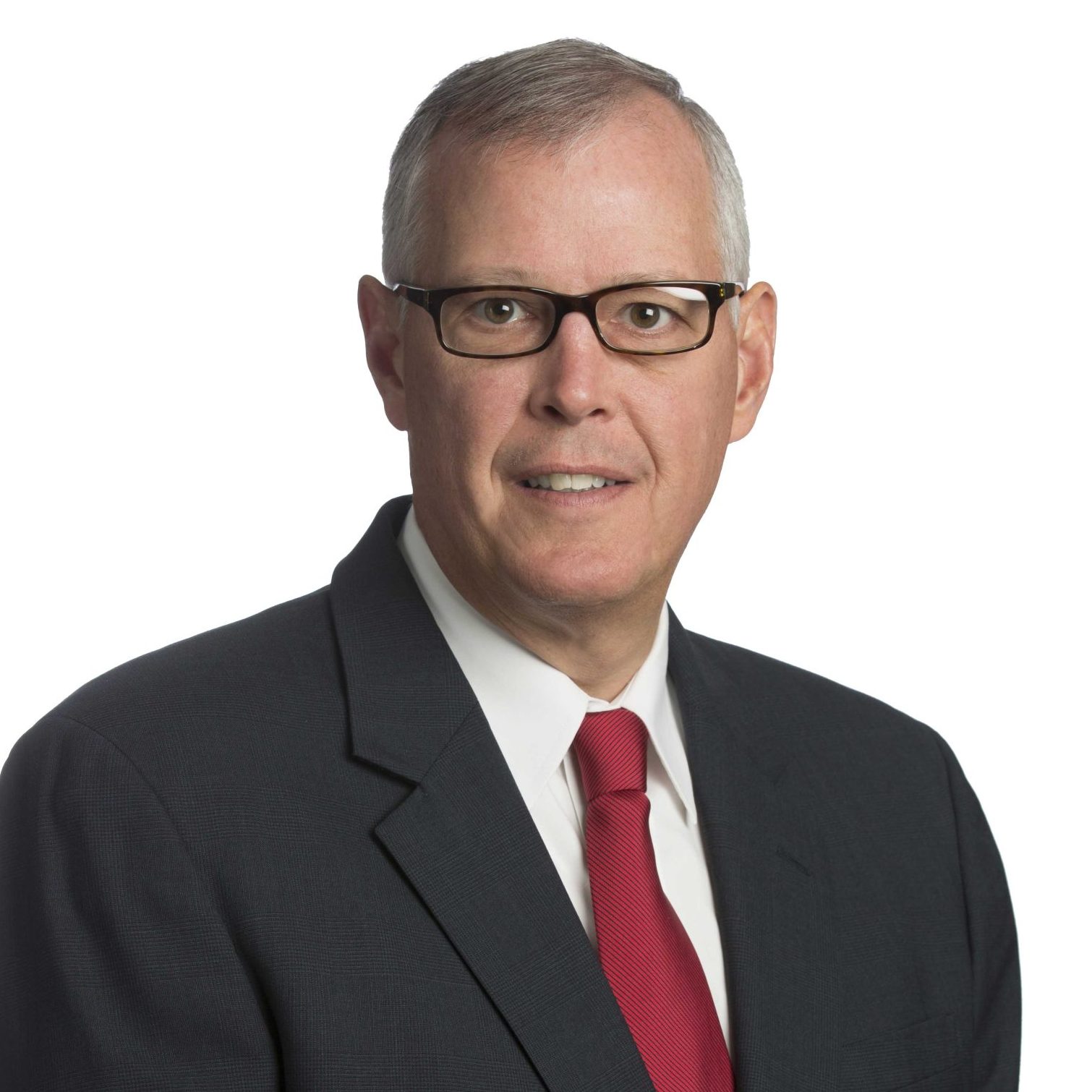 A man wearing glasses, a dark suit, white shirt, and red tie stands against a plain white background.