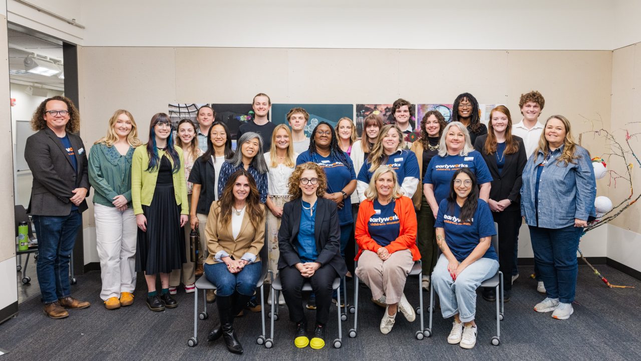 A group of 23 adults, some seated and some standing, pose together indoors in a brightly lit room. Most are smiling and dressed in casual or business casual attire.