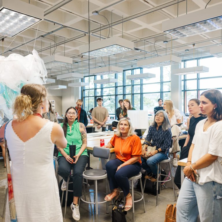 A group of people sit and stand around a woman speaking in a bright, modern classroom with large windows and art installations.