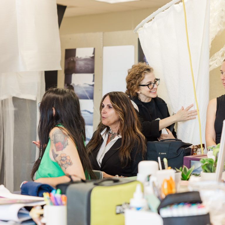 Four women are gathered at a cluttered table in a bright room, engaged in conversation, with art supplies and fabric structures in the background.