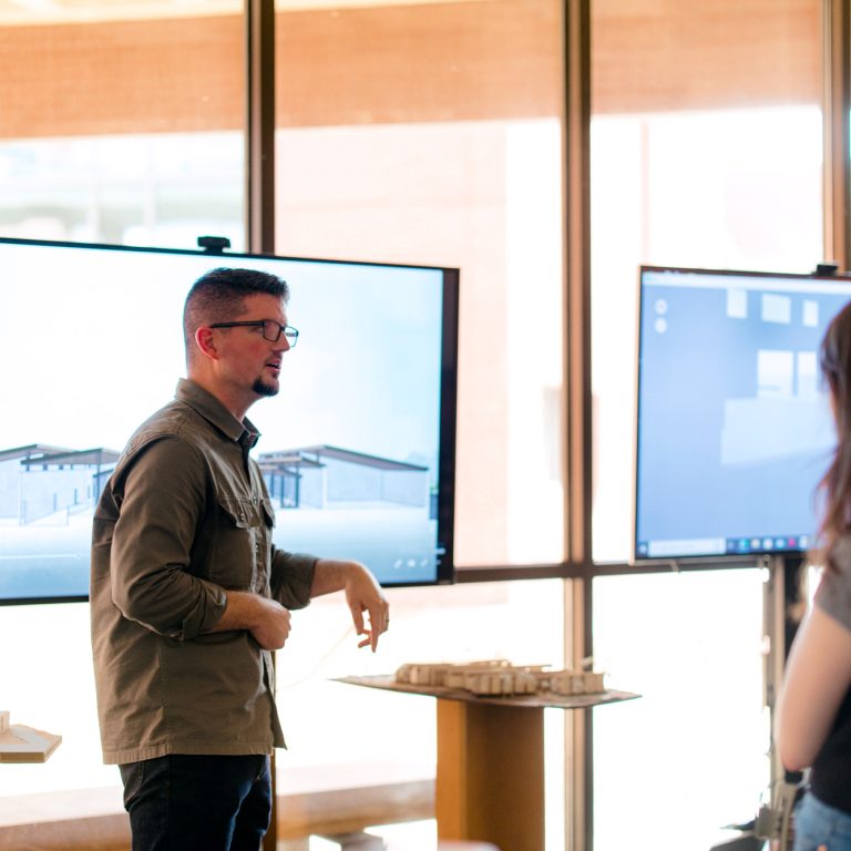 A man stands and gestures while presenting architectural designs on screens to a woman in a well-lit room.