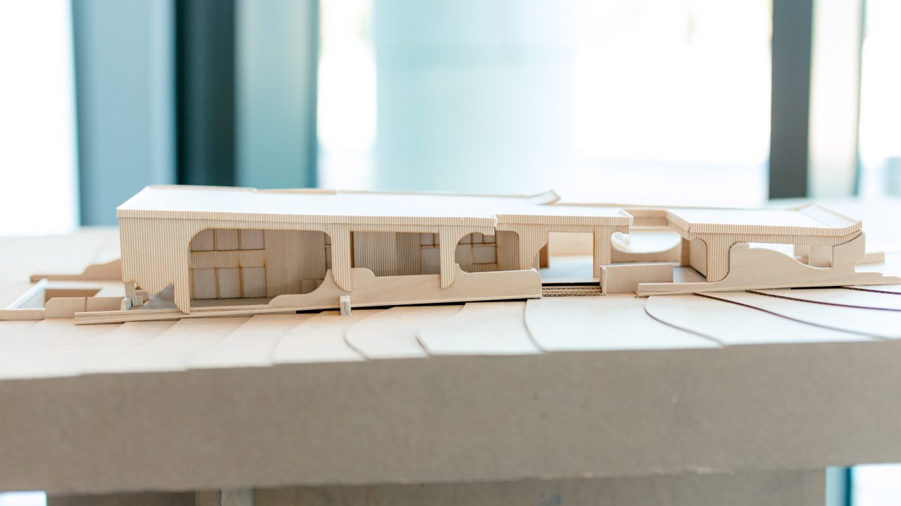 A wooden architectural model displays a modern, elongated building with curved and rectangular cutouts, set on a contoured base.