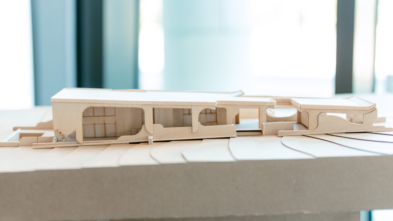 A wooden architectural model displays a modern, elongated building with curved and rectangular cutouts, set on a contoured base.
