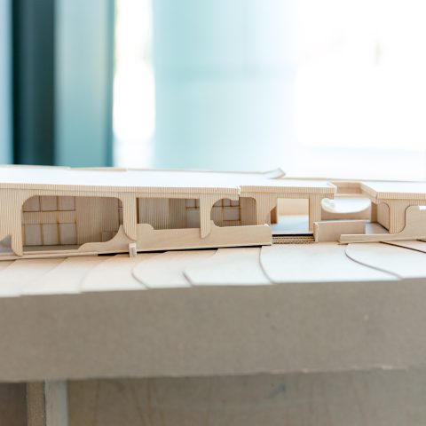 A wooden architectural model displays a modern, elongated building with curved and rectangular cutouts, set on a contoured base.