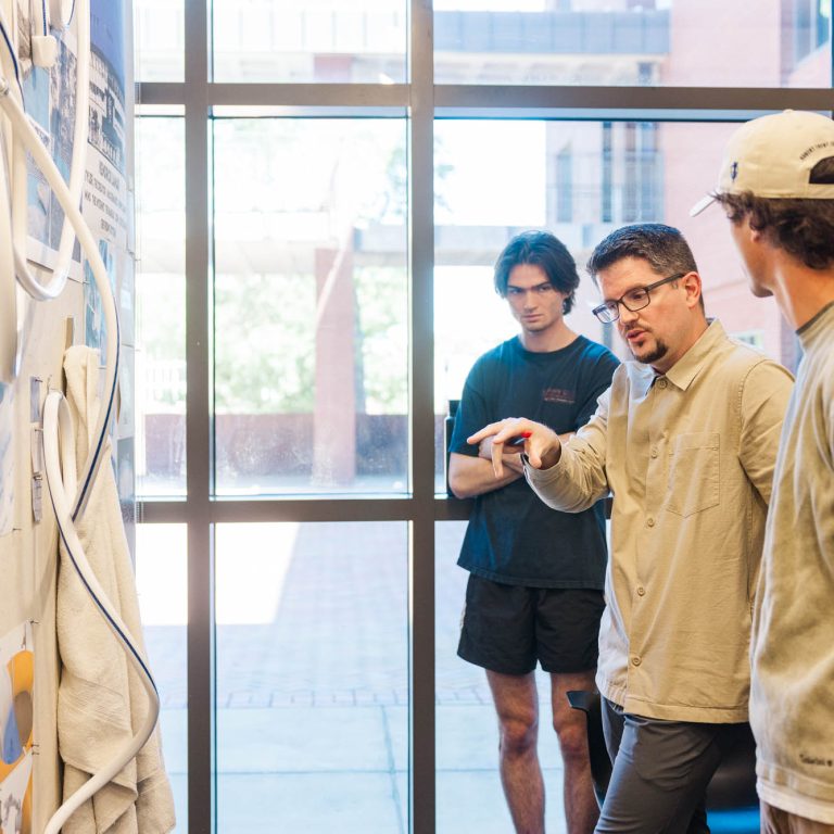 A man gestures and speaks to two people while discussing sketches and plans displayed on a wall in front of a large window.