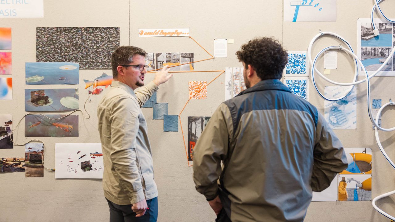 Two men stand in front of a wall covered with photos, diagrams, and notes; one man gestures towards the visuals while explaining something to the other.