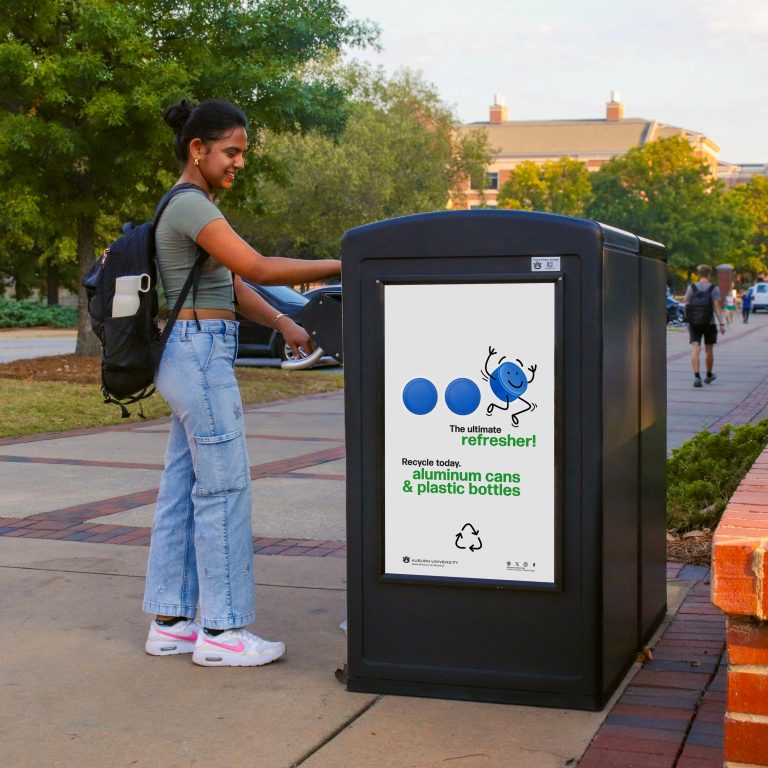 A young woman places a bottle in an outdoor recycling bin with a sign encouraging the recycling of aluminum cans and plastic bottles on a college campus walkway.