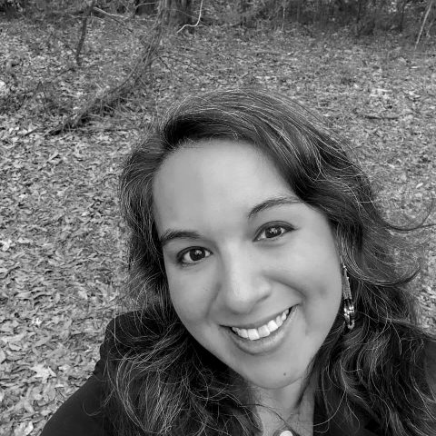 A woman with wavy hair smiles at the camera outdoors in a wooded area with fallen leaves. The photo is black and white.