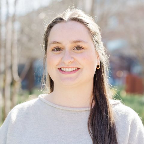 A person with long brown hair and a light gray sweater stands outdoors, smiling at the camera, with trees and buildings in the background.