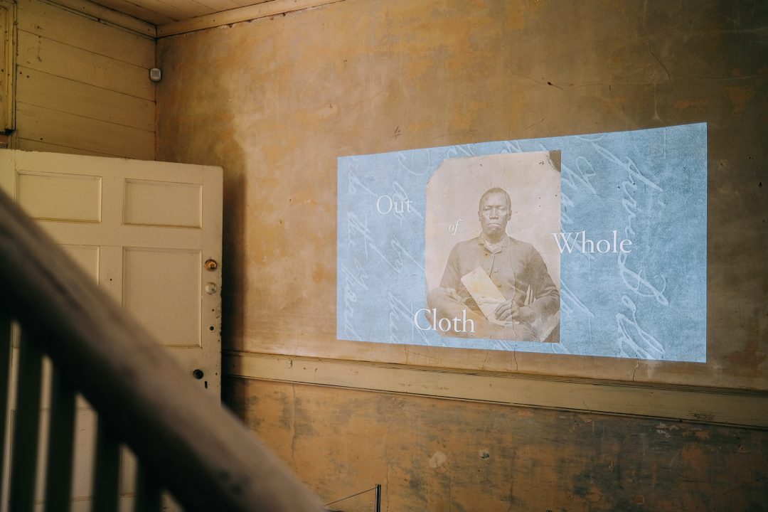 A photograph of a projected image on a worn interior wall, displaying an old portrait of a person with the text Out of Whole Cloth beside it.