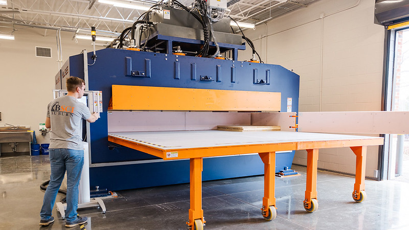 A man operates a large industrial machine with an orange rolling table in a well-lit workshop.