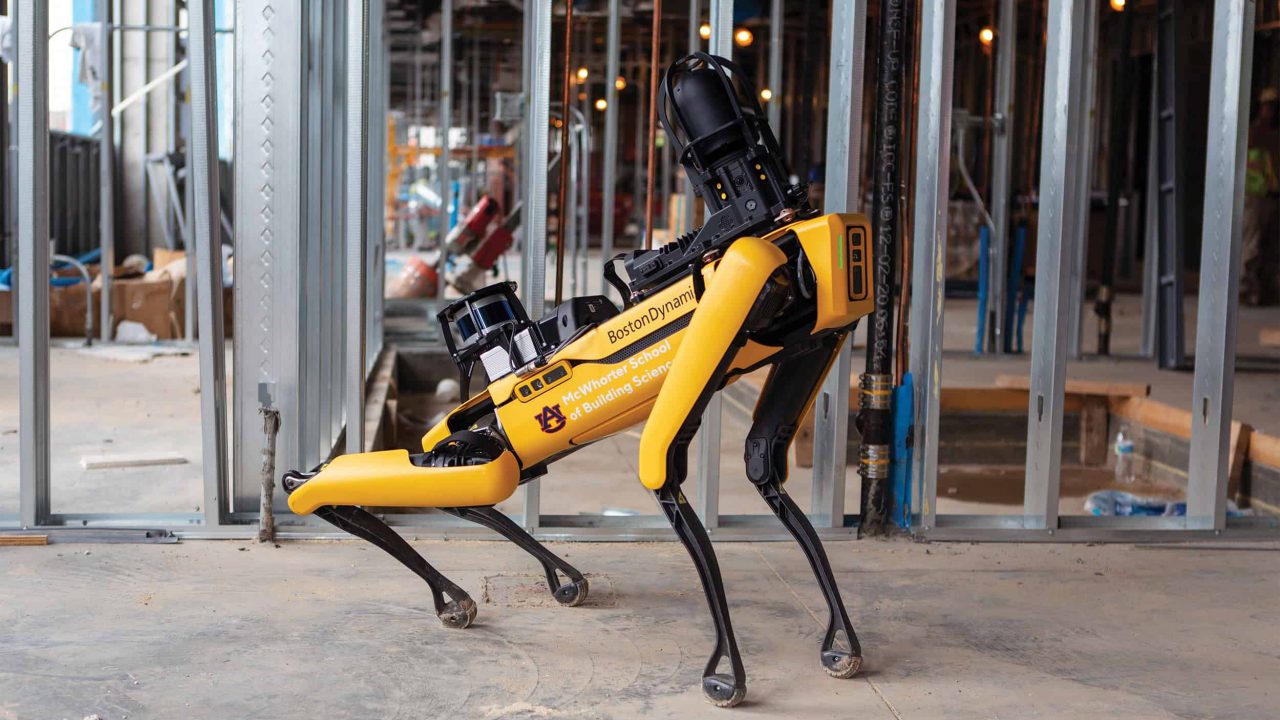 A yellow, four-legged Boston Dynamics robot stands on a construction site with metal framing and various building materials in the background.