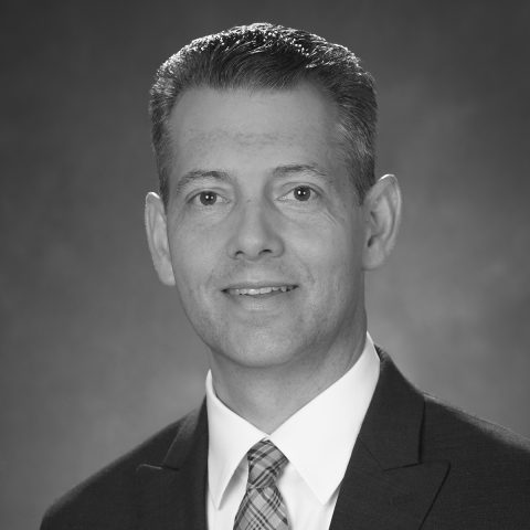 Black and white portrait of a man in a suit and tie, facing the camera and smiling slightly, with a plain background.