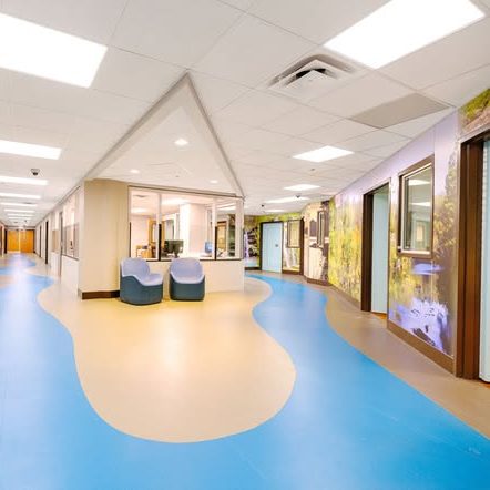 A brightly lit hospital hallway with blue and beige flooring, wall murals of nature scenes, and a central nurses station with two blue chairs.