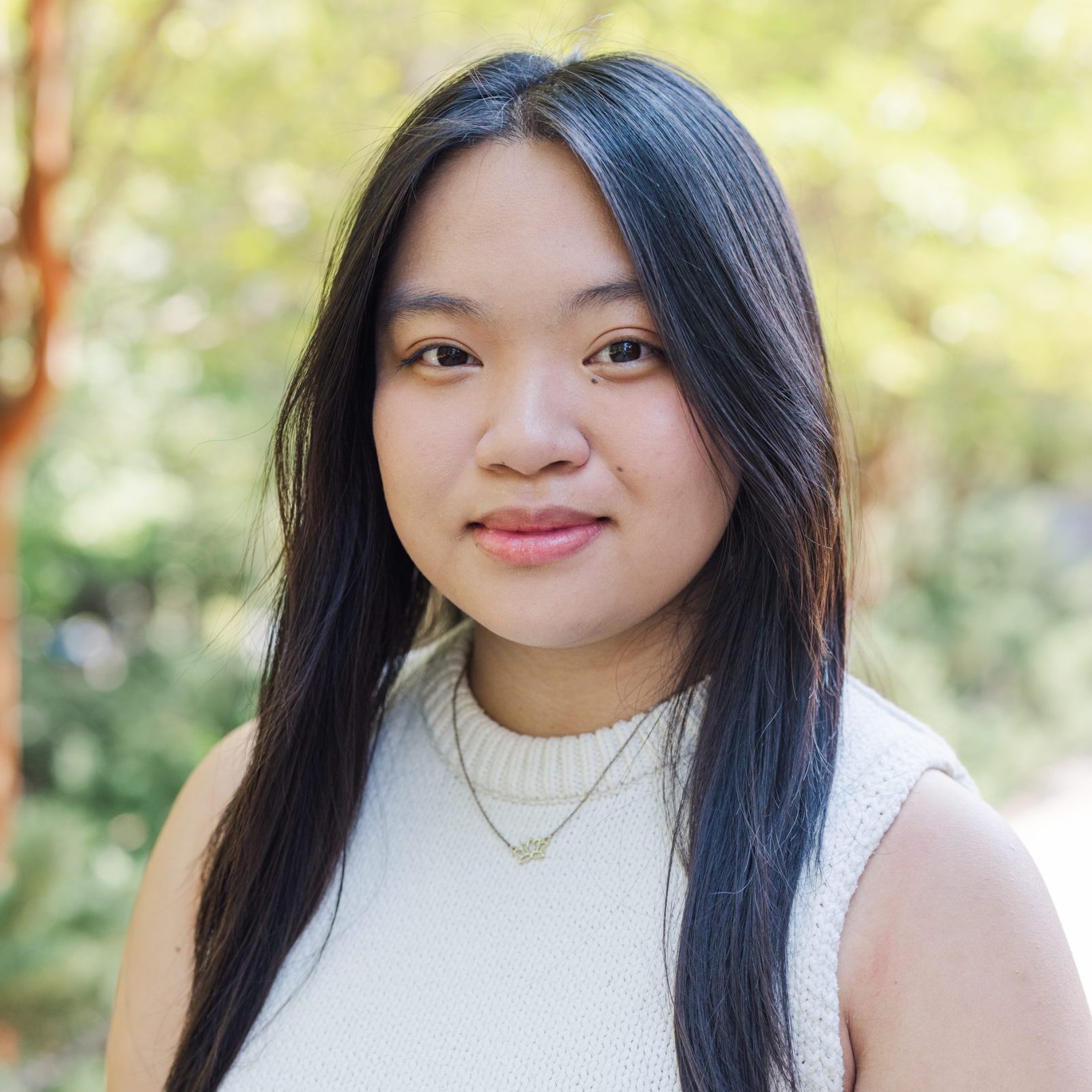 A young woman with long black hair, wearing a white sleeveless top and a necklace, stands outdoors with greenery in the background.