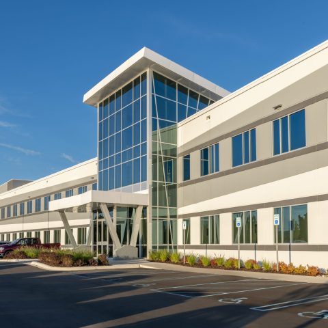 Modern two-story office building with large glass entrance and parking lot, including accessible parking spaces, under a clear blue sky.
