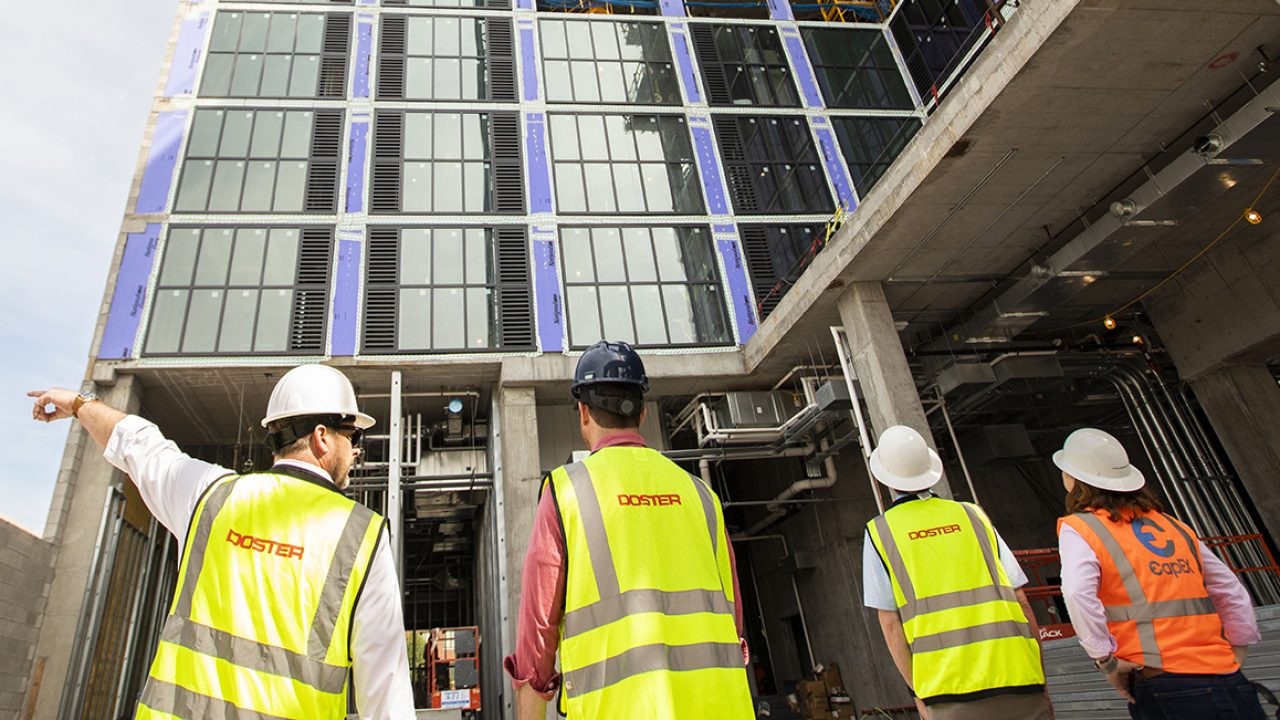 Four construction workers wearing safety vests and helmets observe the exterior of a multi-story building under construction. One worker points toward the structure.