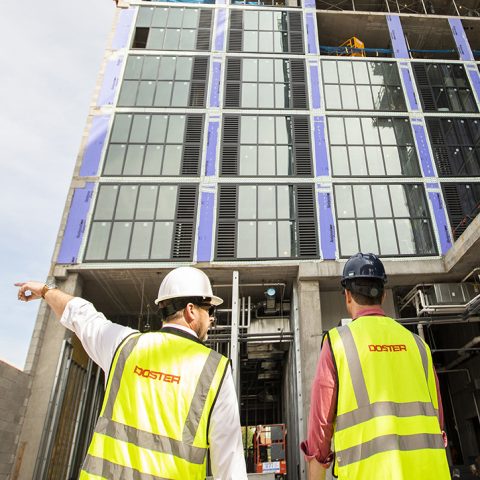 Four construction workers wearing safety vests and helmets observe the exterior of a multi-story building under construction. One worker points toward the structure.