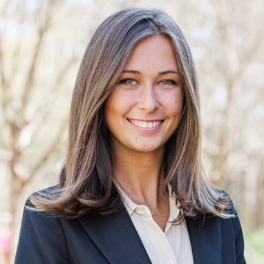 A woman with straight brown hair and a navy blazer over a white blouse stands outdoors, smiling at the camera. Trees and greenery are blurred in the background.
