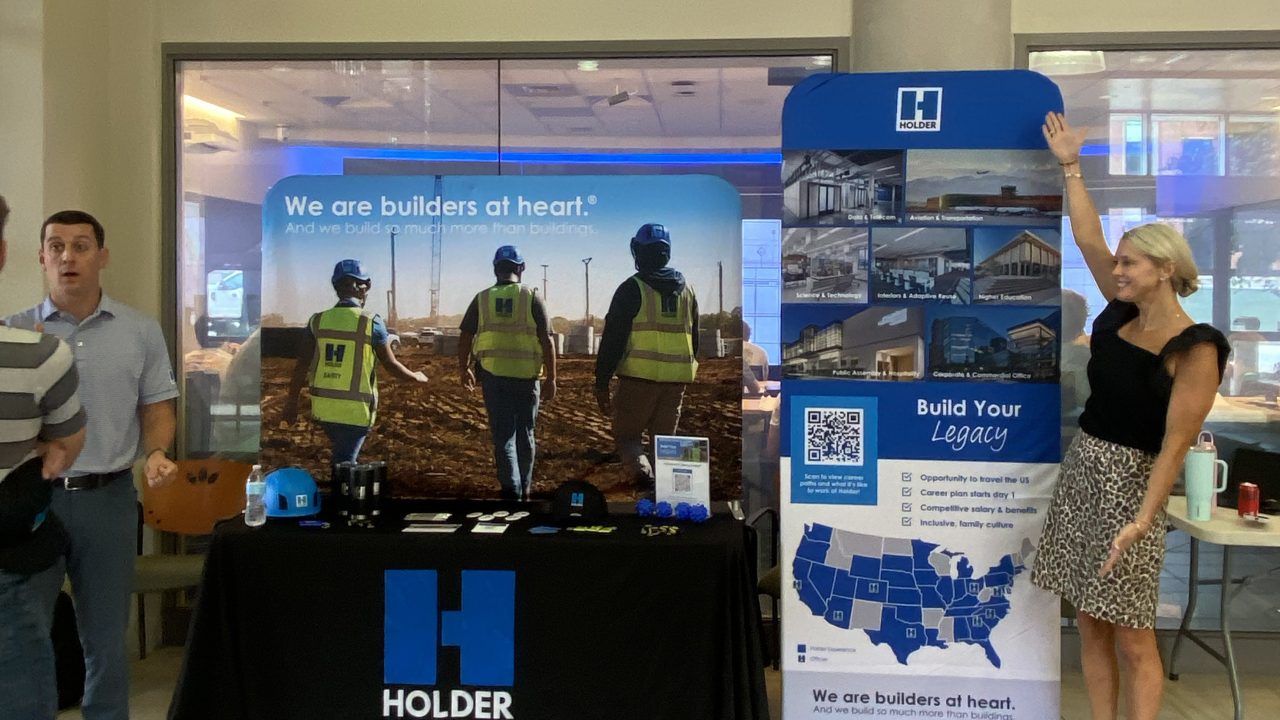 Two people stand at a promotional booth for Holder Construction, featuring banners, informational materials, and branded items on display.