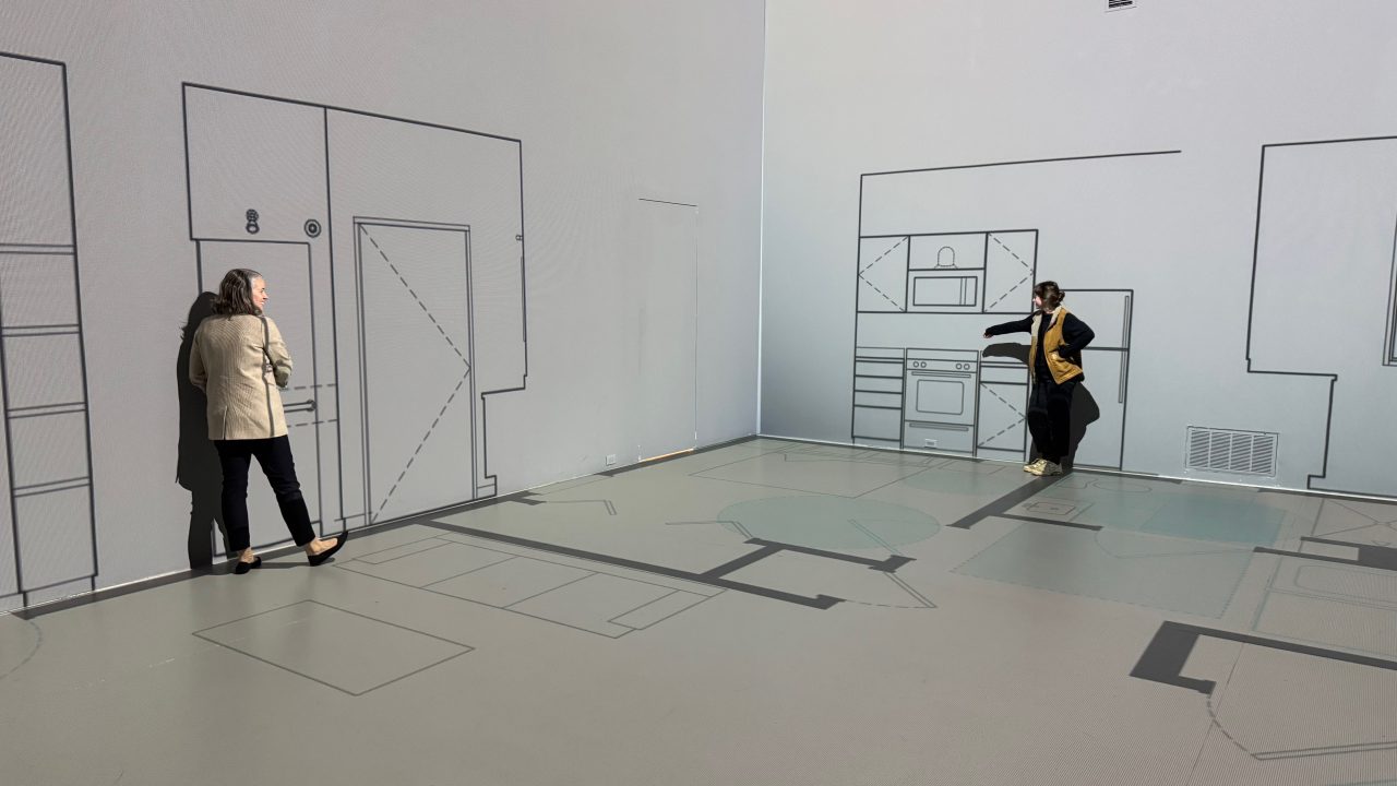 Two people stand in a room with walls and floors decorated with simple black line drawings of kitchen and home interior elements.