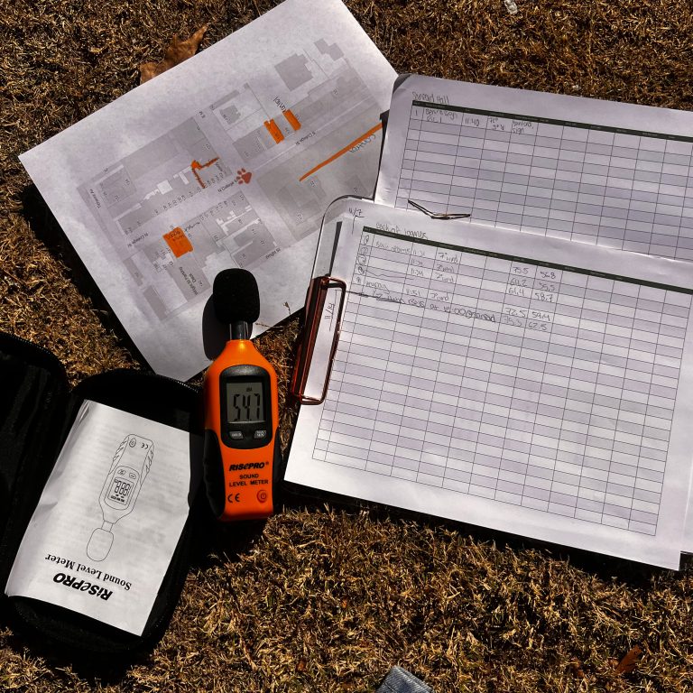 A sound level meter, data sheets with noise measurements, a map, and a carrying case are laid out on dry grass in sunlight.