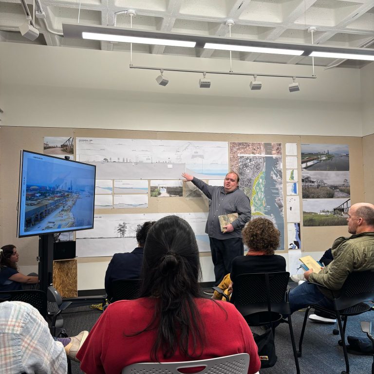 A man stands at the front of a classroom, pointing to design plans on the wall, while several people seated face him and a monitor displaying related images.