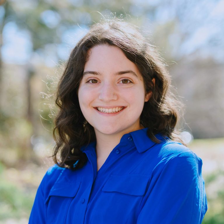 A person with wavy brown hair smiles at the camera, wearing a bright blue button-up shirt outdoors with blurred greenery in the background.