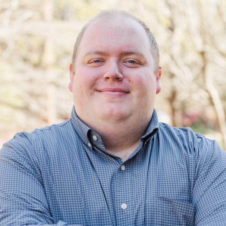 A person wearing a blue checkered shirt stands outdoors with arms crossed, smiling at the camera. Trees without leaves are visible in the background.