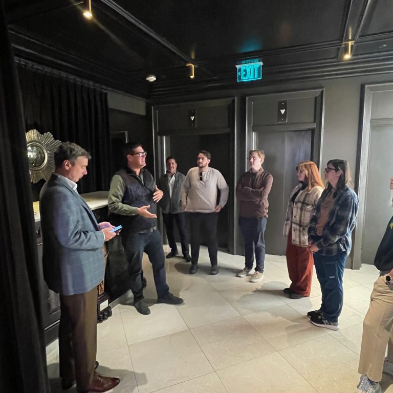 A group of eight people stands in a hallway with dark walls and bright ceiling lights, as one person speaks while others listen attentively.