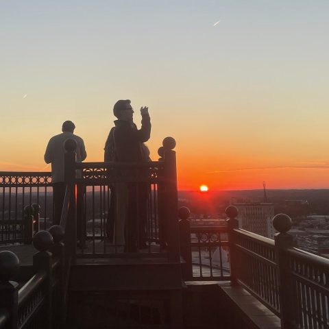 Two people stand on a balcony at sunset; one is taking a photo with a phone while the sun sets over a distant cityscape.