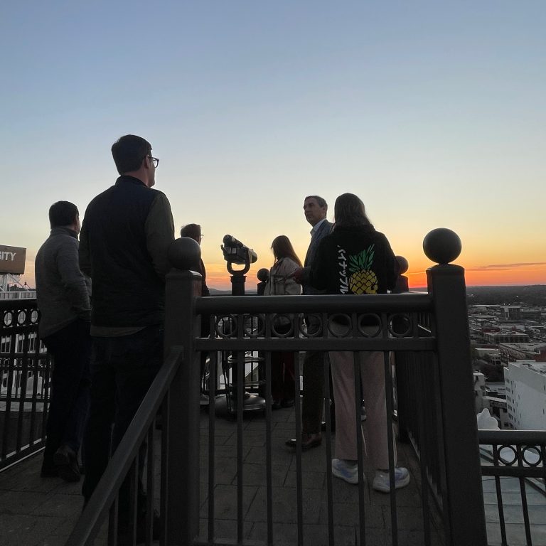 A group of people stand on a rooftop observation deck at sunset, with a cityscape and a mounted binocular viewer in the background.