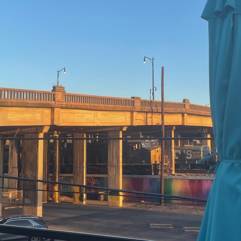 A concrete overpass and lamp posts above a parked train engine and a parking lot, with a rainbow-painted wall in the foreground.