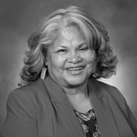 A woman with shoulder-length wavy hair wearing a blazer and patterned top, smiling and posed against a plain studio background.