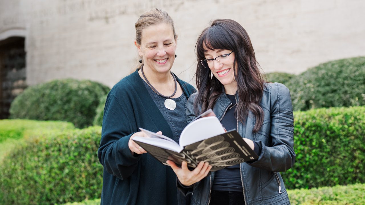 Two women standing outdoors, smiling and looking at an open book together, with green bushes in the background.