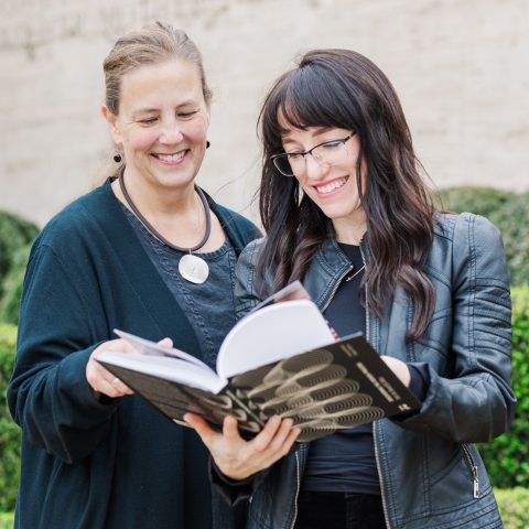 Two women standing outdoors, smiling and looking at an open book together, with green bushes in the background.