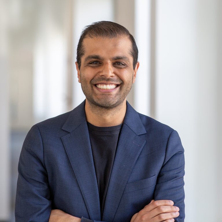 A man with dark hair wearing a blue blazer and black shirt stands smiling with arms crossed in a bright, modern indoor setting.