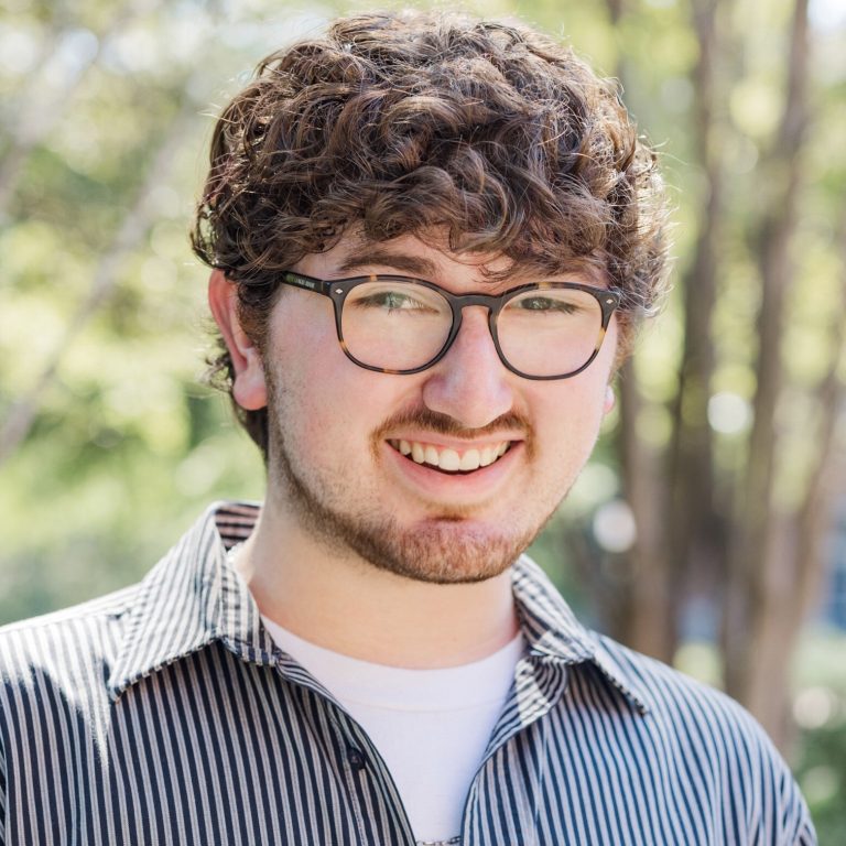 A young man with curly brown hair, glasses, and a short beard smiles outdoors. He is wearing a striped shirt over a white t-shirt. Trees are visible in the background.