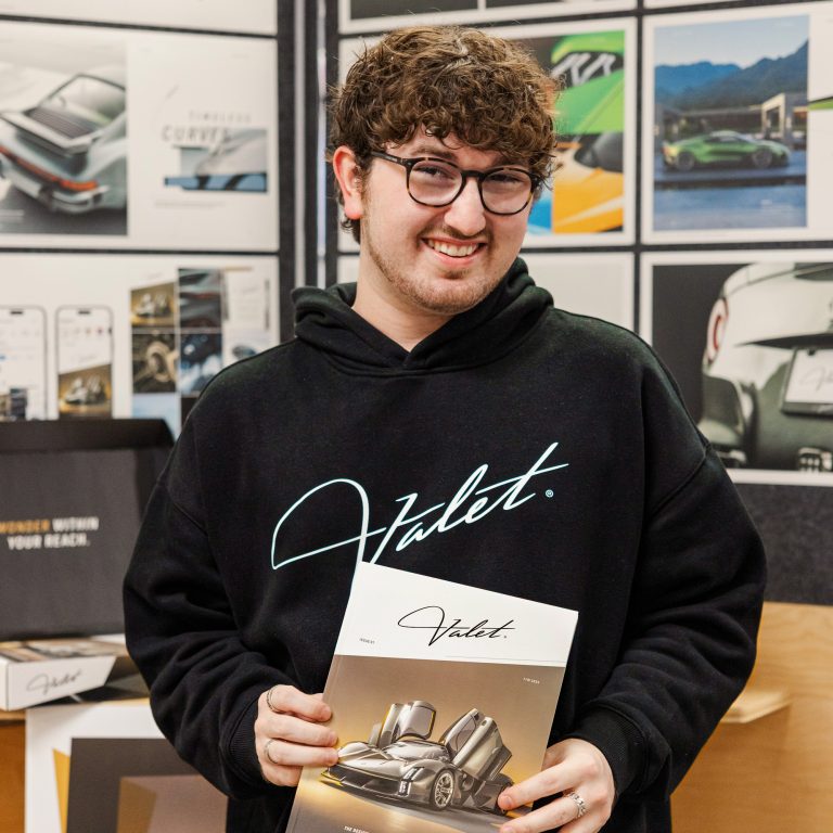 A person wearing glasses and a black Valet hoodie holds a Valet magazine, standing in front of a display featuring car-themed posters and design materials.