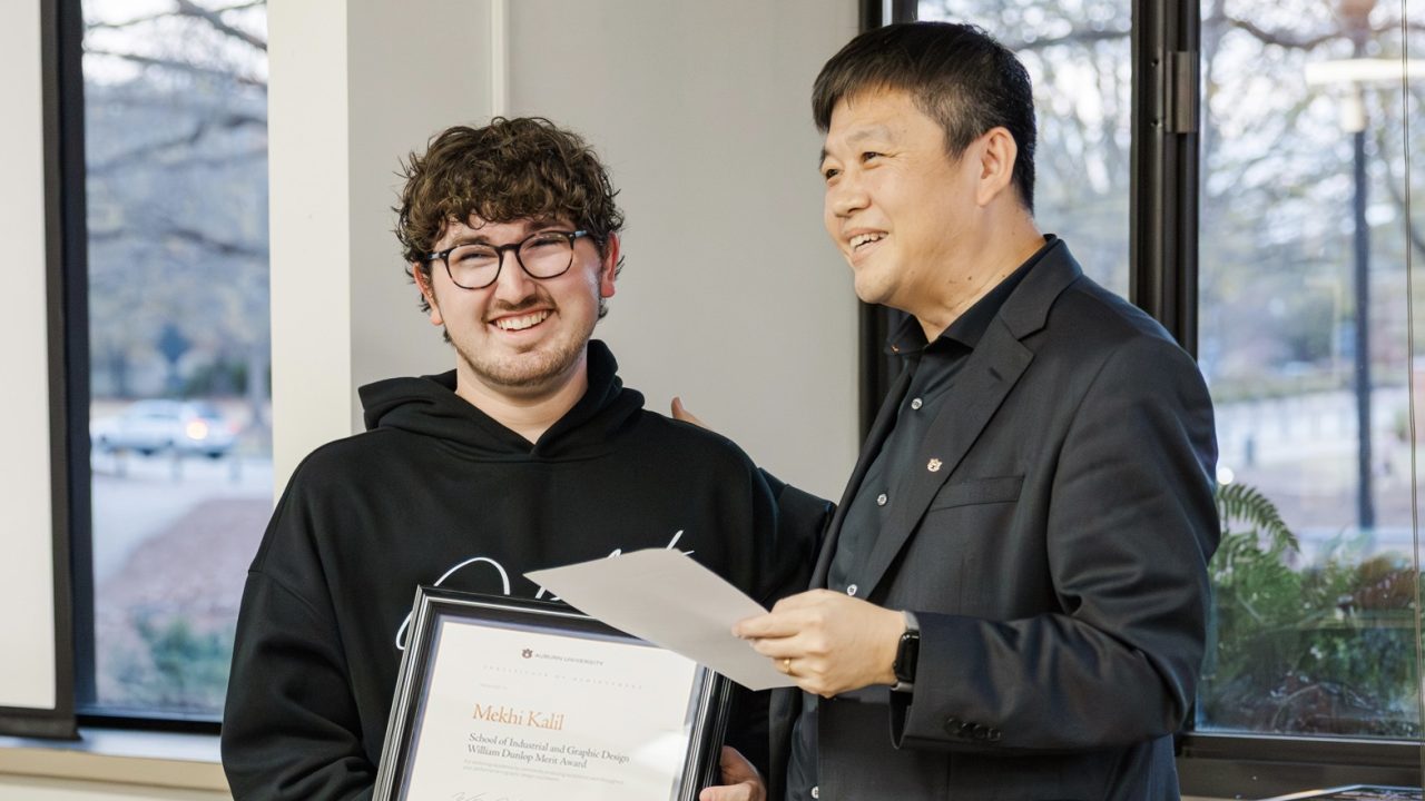 Two people stand indoors by a window. One is holding a framed certificate and smiling, while the other hands over a paper and looks on with a smile.