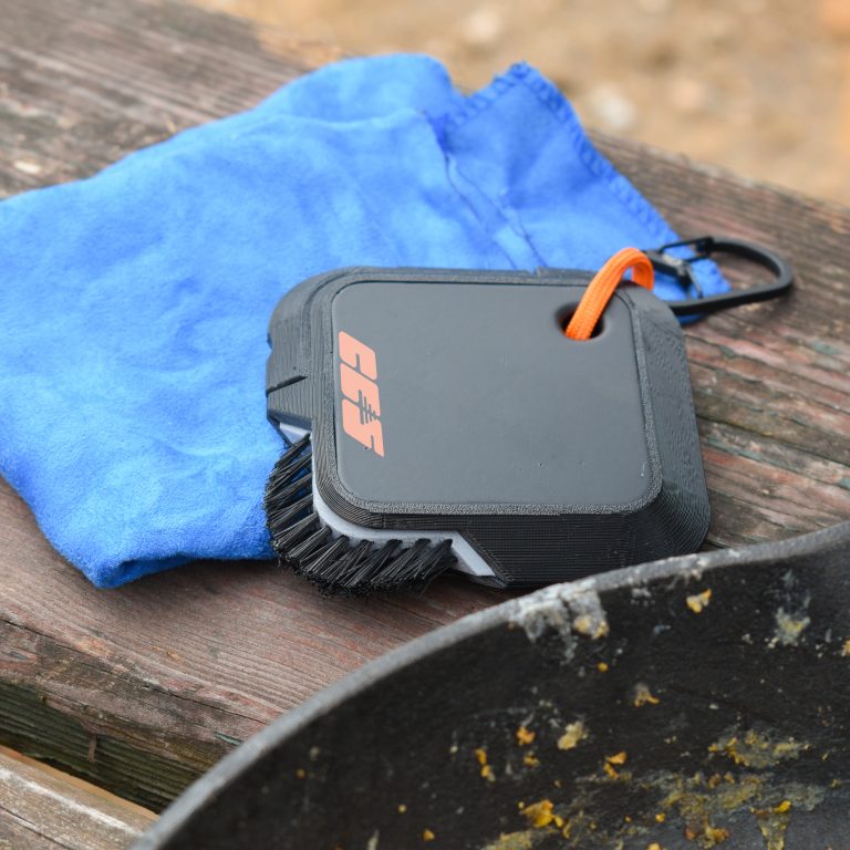 A small cleaning brush and a blue cloth rest on a wooden surface next to a dirty cast iron skillet.