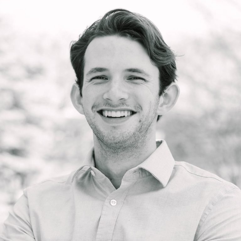 A man with dark hair, wearing a light-colored button-up shirt, stands outdoors with his arms crossed and smiles at the camera.