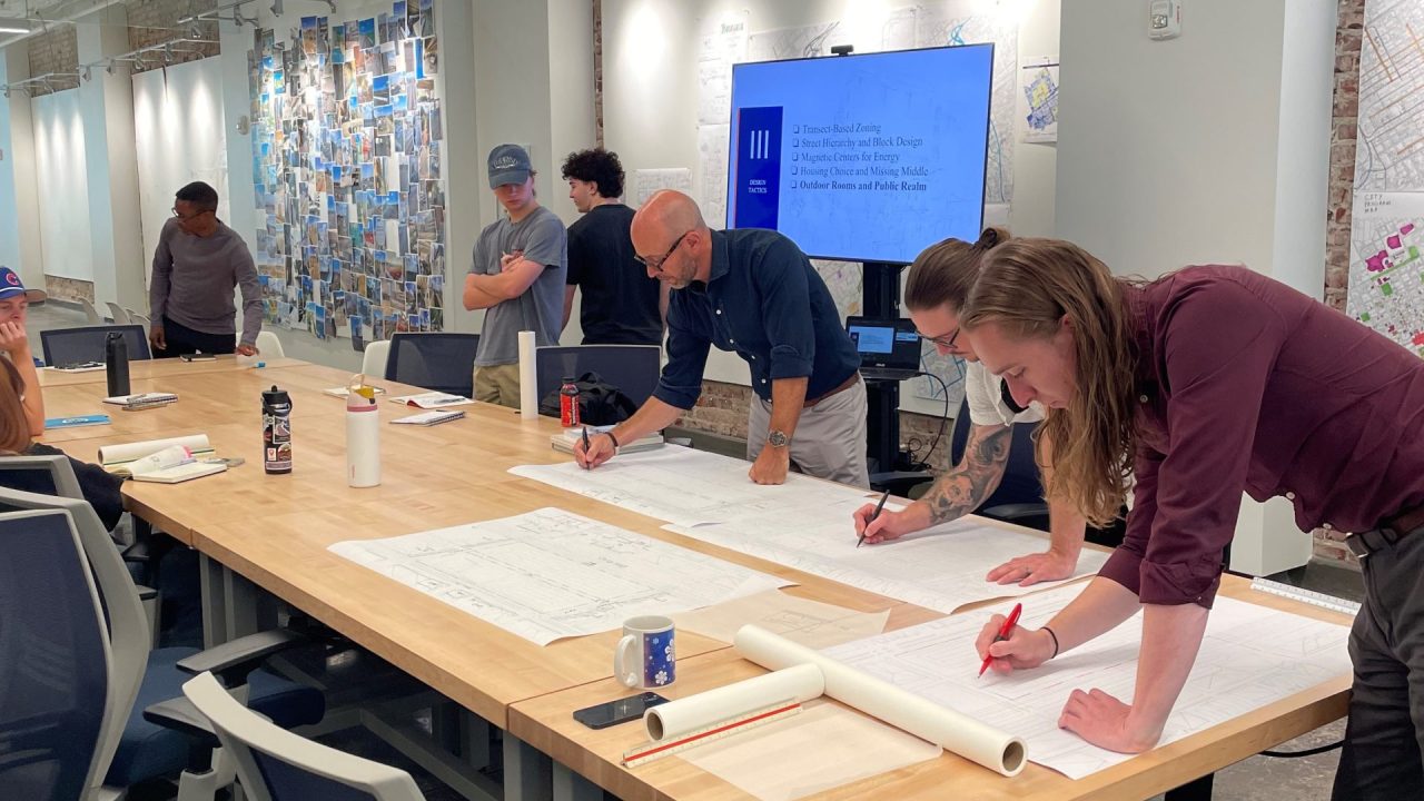 Several people gather around a large table reviewing and marking up architectural plans in a modern design studio.