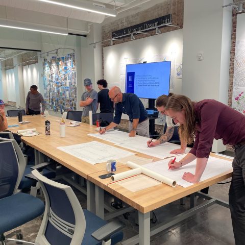 Several people gather around a large table reviewing and marking up architectural plans in a modern design studio.