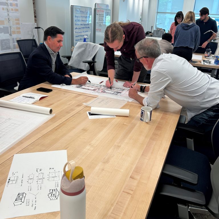 Three people review and discuss architectural drawings on a large table in a modern office; others converse in the background.